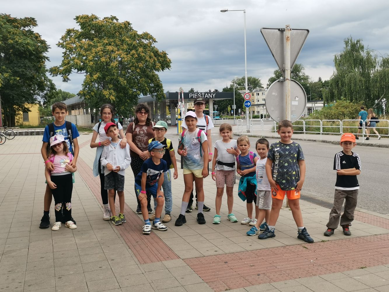 Na fotografii je skupina detí rôzneho veku a dospelých. Všetci stoja na chodníku, okolo nich sú stromy, v pozadí vidieť nápis Piešťany.