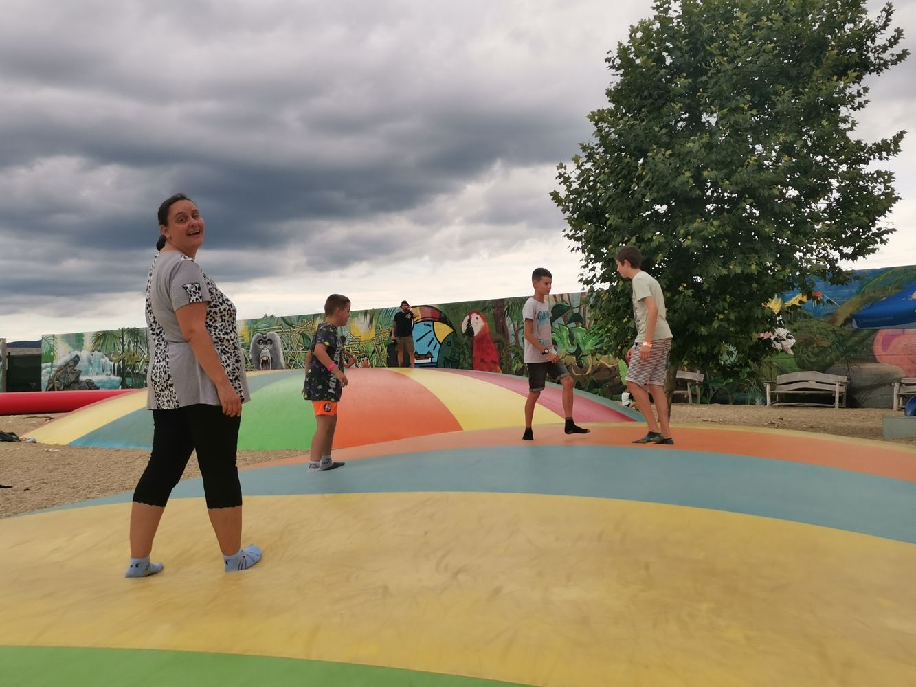 Na fotografii je usmiata žena v sivom tričku a čiernych nohaviciach. Vedľa nej sú traja chlapci, ktorí sa hrajú na trampolíne. V pozadí vidieť veľký strom a stenu s farebnou maľbou rôznych zvierat a prírodnými motívmi.