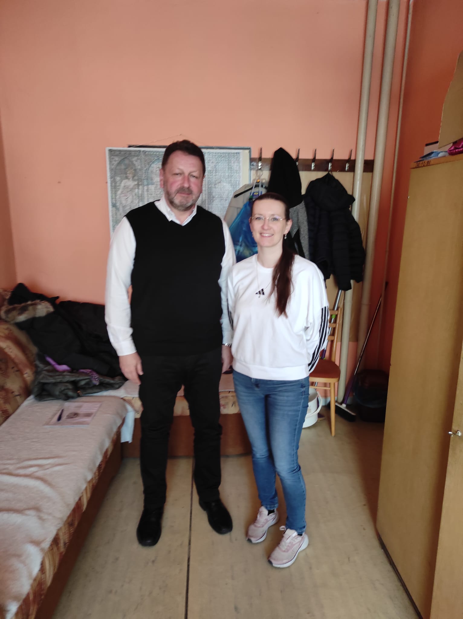Na fotografii sú dve osoby. Muž vľavo má krátke tmavé vlasy a bradu; má na sebe bielu košeľu, čiernu pletenú vestu, čierne nohavice a čierne topánky. Žena vpravo má dlhé tmavé vlasy zopnuté do chvosta a okuliare; má na sebe bielu mikinu s čiernymi pruhmi na rukávoch, modré rifle a svetloružové tenisky.