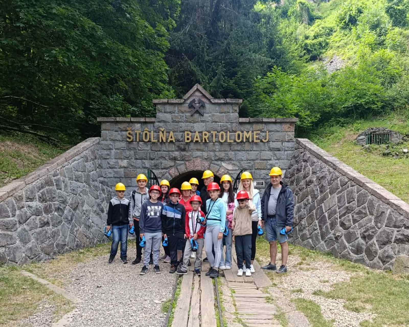 Na kamennom portáli je veľký nápis: „ŠTÔLŇA BARTOLOMEJ“ a nad ním je symbol dvoch skrížených baníckych kladív. Pred vstupom stojí skupina detí a dospelých v ochranných prilbách, väčšinou žltých, niektoré sú červené. Majú na sebe bežné športové a turistické oblečenie, niektorí držia modré baterky alebo lampy. Vchod do štôlne je oblúkový, z tmavej kamennej steny, z oboch strán vedú šikmé kamenné múry. Pred nimi sú koľajnice, ktoré smerujú k vstupu. Okolie tvorí svah pokrytý trávou a hustým zeleným lesom v pozadí.
