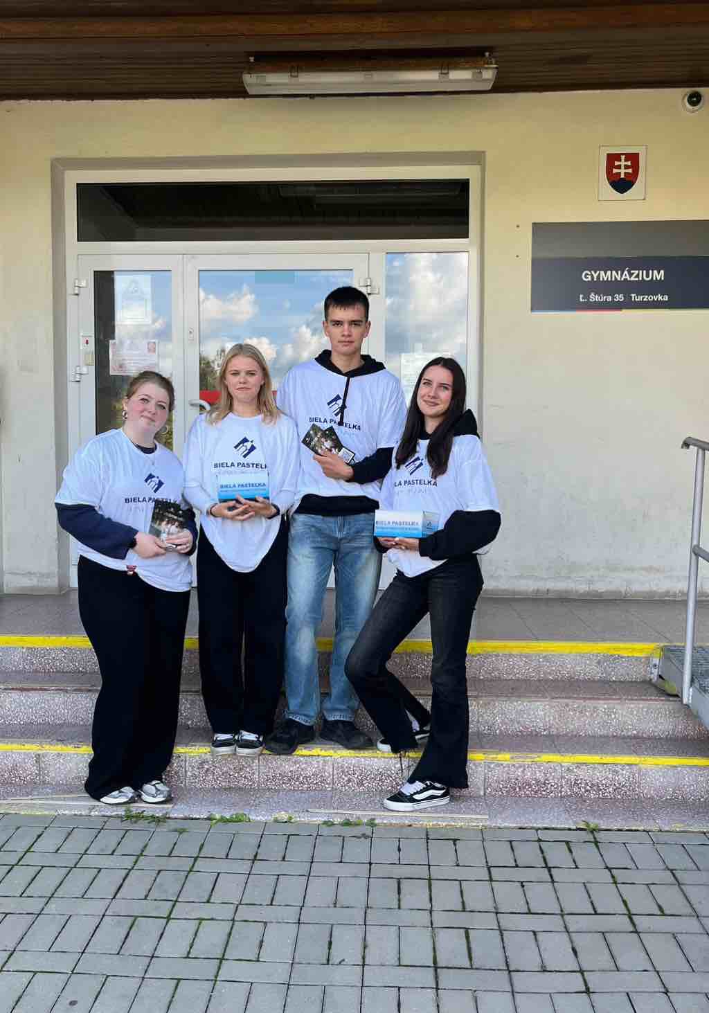 Štyria mladí ľudia stoja na schodoch pred vstupom do budovy. Majú oblečené biele tričká s nápisom „BIELA PASTELKA“ a logom zloženým z dvoch šikmých paličiek a bodky. Dve dievčatá držia v rukách pokladničky – biele škatule s modrým spodným pásom a textom „BIELA PASTELKA“. Chlapec a dievča držia malé letáky . Na pravej strane steny je tabuľa s nápisom „GYMNÁZIUM“ a pod ním „Ľ. Štúra 35 Turzovka“. Nad tabuľou je štít so slovenským štátnym znakom.