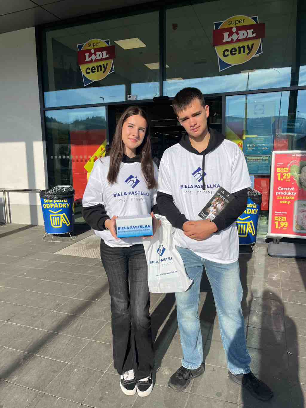 Dvojica mladých ľudí v bielych tričká s modrým logom a textom „Biela pastelka“. stojí pred vchodom do predajne Lidl. Dievča vľavo má dlhé hnedé vlasy, čiernu mikinu pod tričkom, tmavé nohavice a čierno-biele tenisky. V rukách drží modrú pokladničku s nápisom „BIELA PASTELKA“. Chlapec vpravo má krátke hnedé vlasy, čiernu mikinu pod tričkom, svetlomodré rifle a čierne topánky. V jednej ruke drží letáky, v druhej bielu plastovú tašku s logom „Biela pastelka“. Za nimi sú veľké presklené dvere a okná vstupu do Lidla s viacerými reklamnými plagátmi a nápismi „super Lidl ceny“. Na oboch stranách dverí sú modré nádoby na odpadky so žltým symbolom koša. Slnko svieti z pravej strany, postavy vrhajú dlhé tiene na dlažbu pred obchodom.