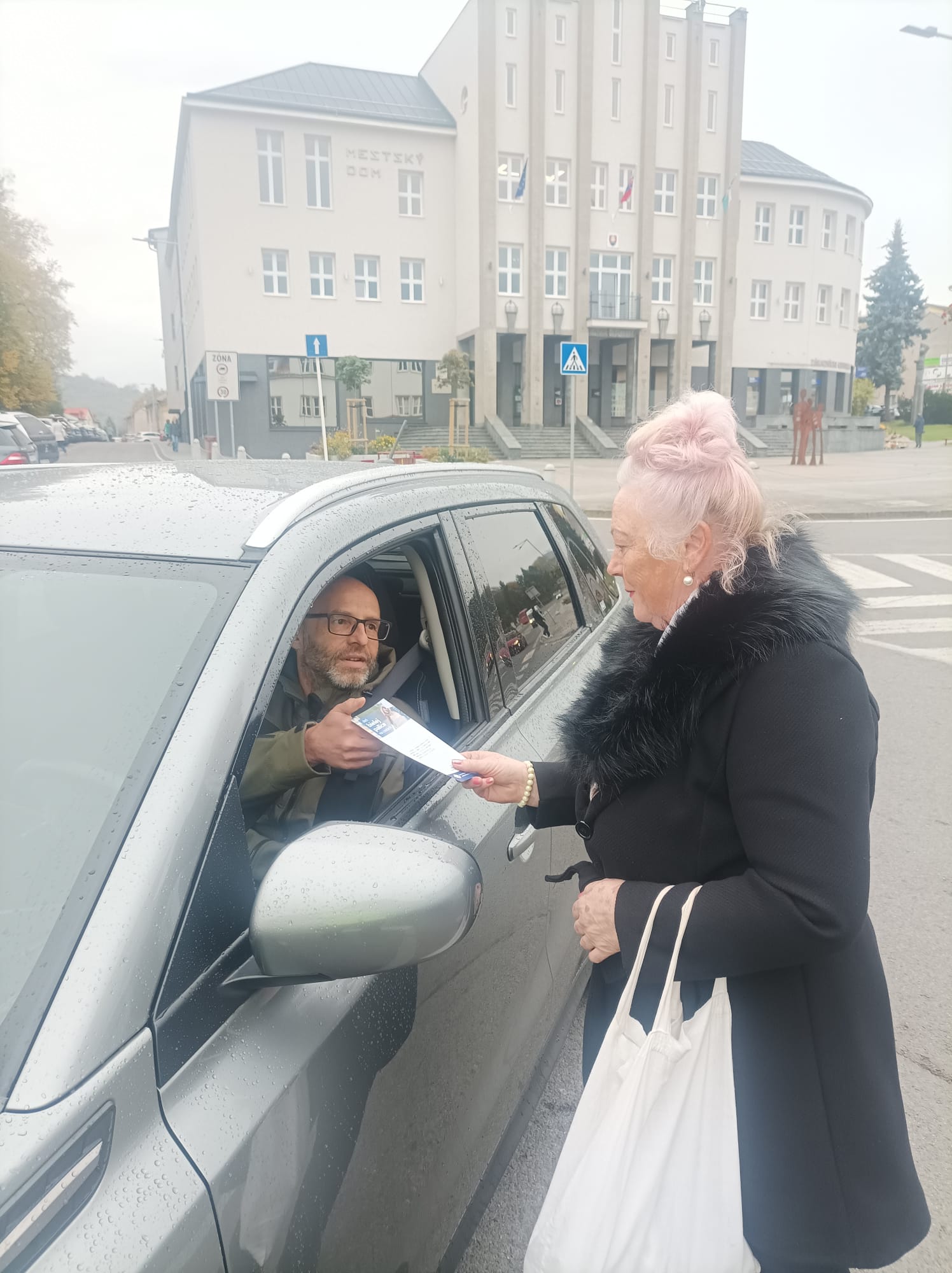 Žena so svetloružovými vlasmi v elegantnom čiernom kabáte stojí pri striebornom aute a podáva leták mužovi sediacemu na mieste vodiča so stiahnutým oknom. Muž si leták preberá. V pozadí je väčšia svetlosivá budova s nápisom „MESTSKÝ DOM“, Pred budovou je priechod pre chodcov so zvislou dopravnou značkou a časť križovatky.