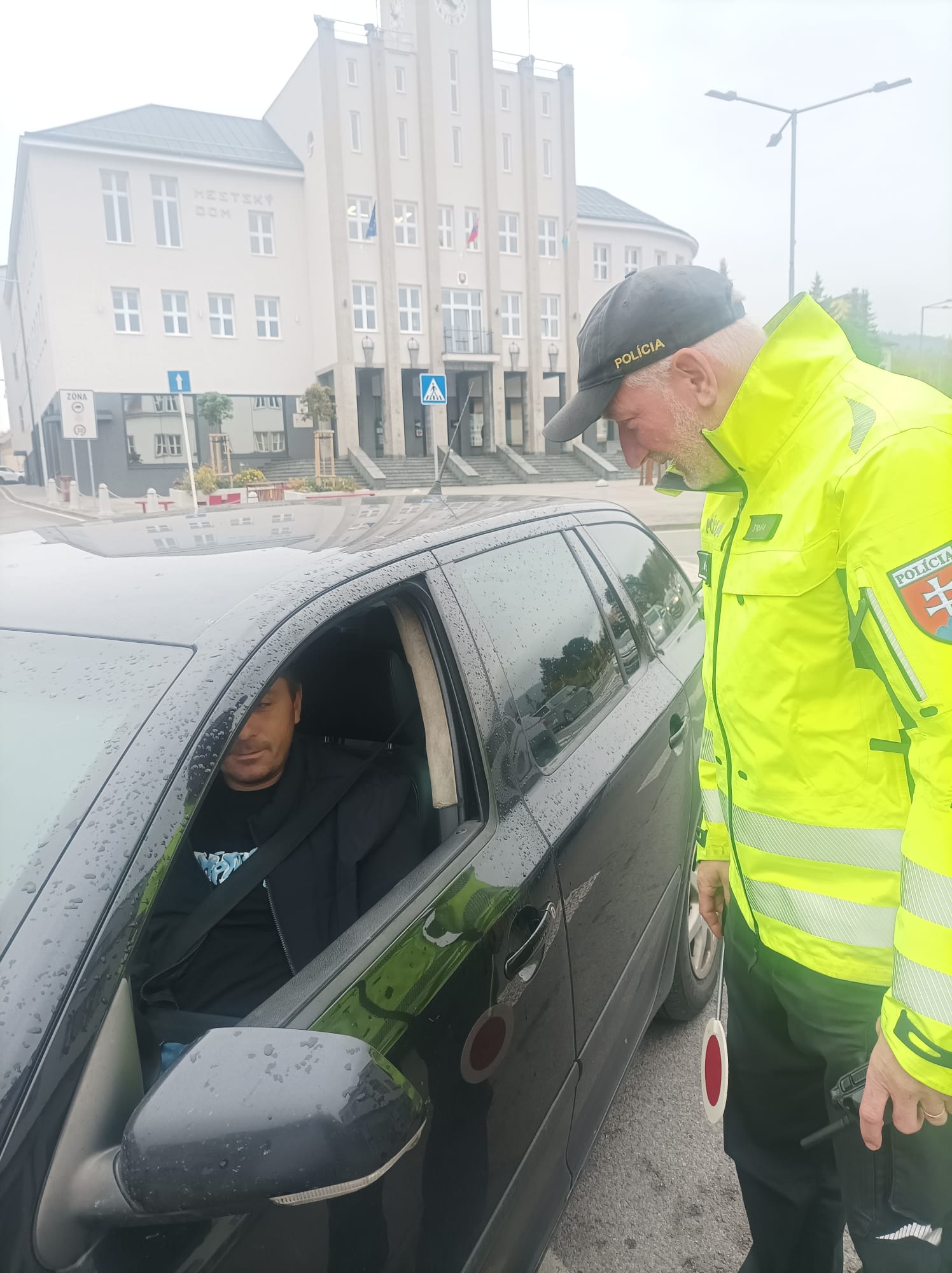Policajt v reflexnej žltej bunde a čiernej šiltovke s nápisom „POLÍCIA“ stojí pri čiernom aute pri okne vodiča, v ruke má bielo‑červenú terčovku. Vodič sedí vpredu a má zapnutý pás. Okno je stiahnuté. Auto je mokré od dažďa.